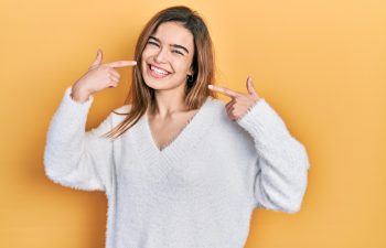 A smiling young woman with brown hair, wearing a white fuzzy V-neck sweater, points to her wide, happy smile with both index fingers. The background is a solid yellow/orange color.