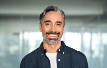 A headshot of a handsome, middle-aged man with salt-and-pepper hair and a beard, smiling warmly at the camera. He is wearing a dark blue button-down shirt over a white t-shirt, with a blurred modern office environment in the background.