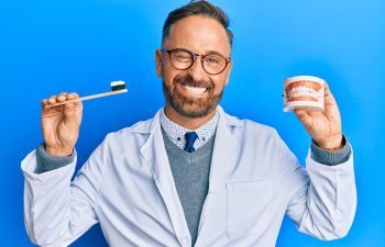 A male dentist with a beard and glasses wearing a white coat, winking while holding a toothbrush and a model of a jaw on a blue background.